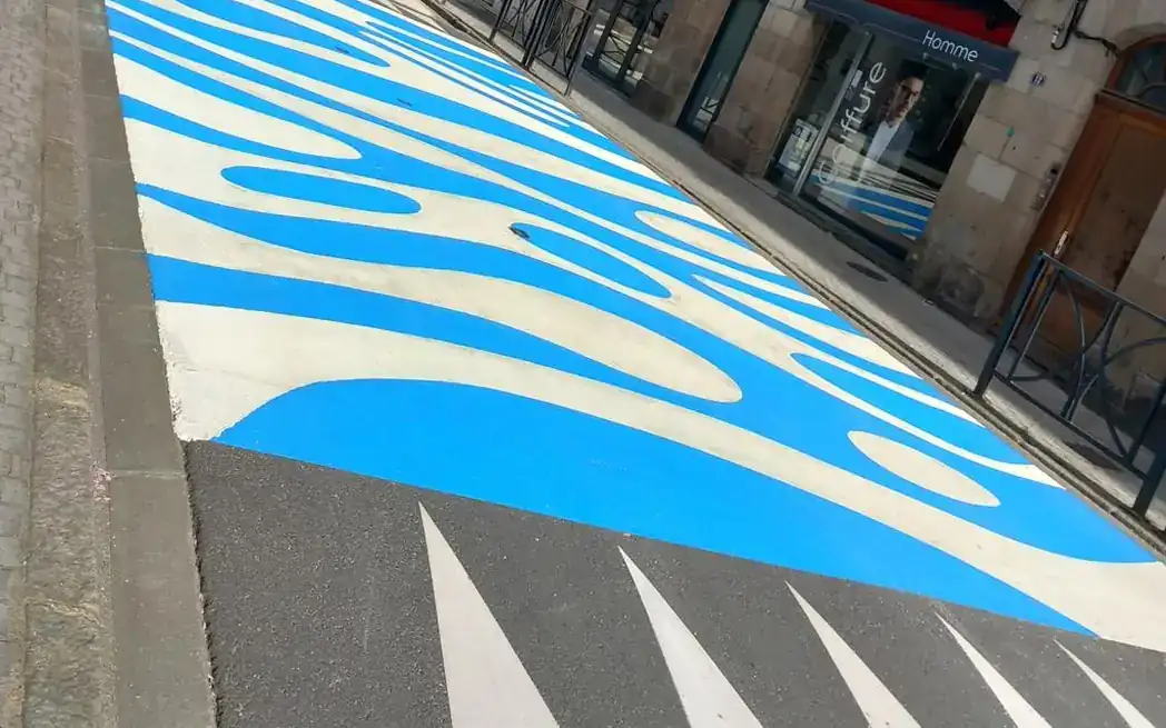 Motif de vagues bleues peint sur la chaussée d’une rue commerçante, précédé de marquages triangulaires blancs